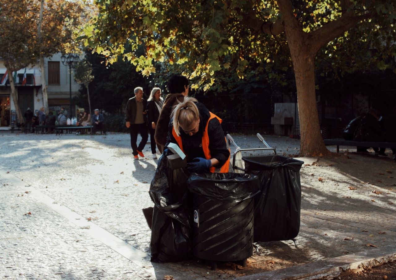 our-story-02 Sanitation worker in orange vest collecting trash in urban park during fall.